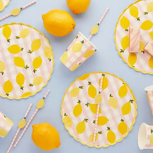 Lemon-themed party tableware with yellow lemon print plates, cups, straws, and whole lemons arranged on a light background